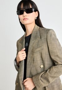 Ruskea ruudullinen bleiseri, jossa on jämäkkä muotoilu, kaarevat laput, musta nappi ja hartiatyynyt, kerroksittain mustan T-paidan päällä.