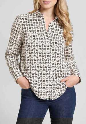 Donna che indossa una blusa color crema con motivi a foglia neri e jeans blu scuro, con le mani in tasca, in piedi davanti a uno sfondo chiaro e uniforme.