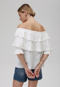 Witte off-shoulder top met drie gelaagde ruffles, gestructureerde stof en korte mouwen, gecombineerd met denim shorts.