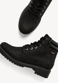 Svarta ankelstövlar tillverkade av läder med matt yta, med metallöglor, tjocka sulor och en vadderad krage. Diskret logodetalj finns.