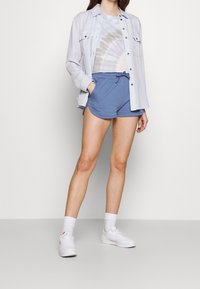 Ljusblå skjorta med knappar över en tie-dye t-shirt, matchad med blå shorts. Vita sneakers och strumpor fulländar outfiten.