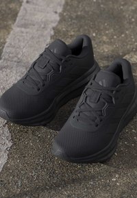 Paire de baskets de sport noires avec une maille texturée et des lacets, posées sur un bitume rugueux près d'une ligne blanche fanée.