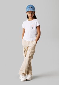 Camiseta blanca de manga corta con bordado de flores, pantalones cargo beige con bolsillos y una gorra de mezclilla azul clara. Zapatillas blancas completan el look.