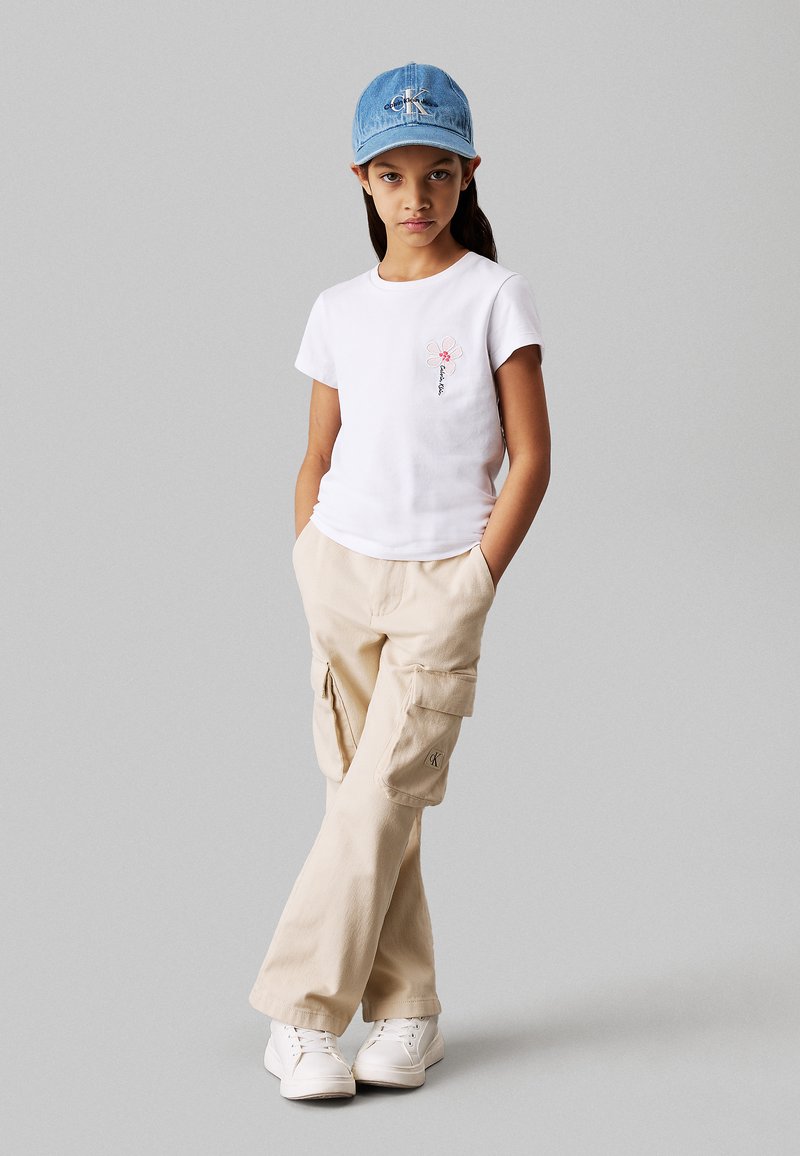Camiseta blanca de manga corta con bordado de flores, pantalones cargo beige con bolsillos y una gorra de mezclilla azul clara. Zapatillas blancas completan el look.