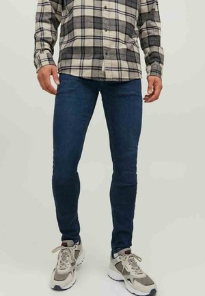 Mężczyzna ubrany w ciemnoniebieskie obcisłe dżinsy, beżową i czarną koszulę w kratę zapinaną na guziki oraz beżowo-białe tenisówki na jednolitym tle.