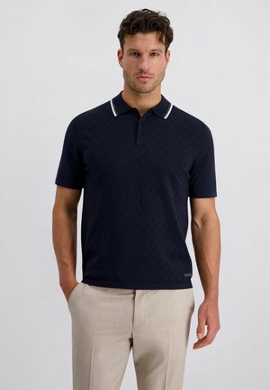 Mann trägt ein marineblaues Polohemd mit kurzen Ärmeln und strukturiertem Muster sowie beige Hosen, steht vor einem einfarbigen hellen Hintergrund.
