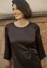 Brun satinblus med trekvartslånga ärmar, rund hals och tvistdetalj vid midjan. Slät textur mot en enkel bakgrund.