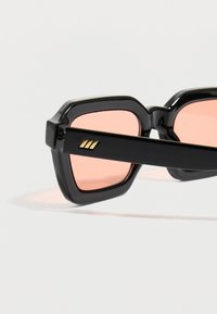 Μαύρα γυαλιά ηλίου με παχιά πλαίσια και ροζ φακούς με απόχρωση, διακοσμημένα με μικρή χρυσή τριπλή γραμμή στο πλάι του βραχίονα, πάνω σε ανοιχτό φόντο.