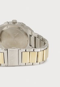 Metallarmbanduhr mit einem Edelstahlband, das goldene Akzente aufweist. Das Zifferblatt ist rund mit minimalistischen Markierungen und einer strukturierten Oberfläche.