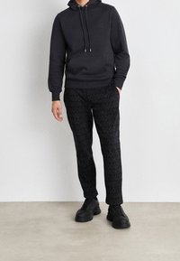 Veste à capuche noire avec une poche avant, associée à un pantalon noir présentant un motif de logo répétitif. Complété par des chaussures noires massives.