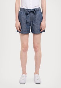Person som bär blå denimshorts med ett knytet midjebälte, vit knappad topp och vita sneakers, som står mot en enkel bakgrund.