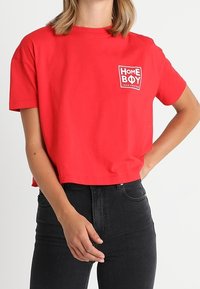 Camiseta corta roja de algodón con mangas cortas. Presenta un parche de logo blanco con la inscripción "HOME BOY" en la parte izquierda del pecho.