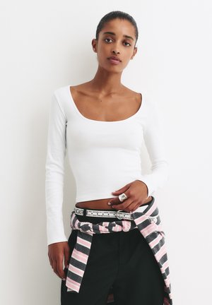 Haut blanc à manches longues en côtelé avec un décolleté rond, associé à un pantalon noir et une chemise enstripée rose et grise nouée en accessoire.