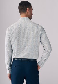 Langærmet skjorte med hvid bund og flerfarvet blomsterprint; glat stof med standardkrave og knap på manchet, kombineret med mørke bukser.