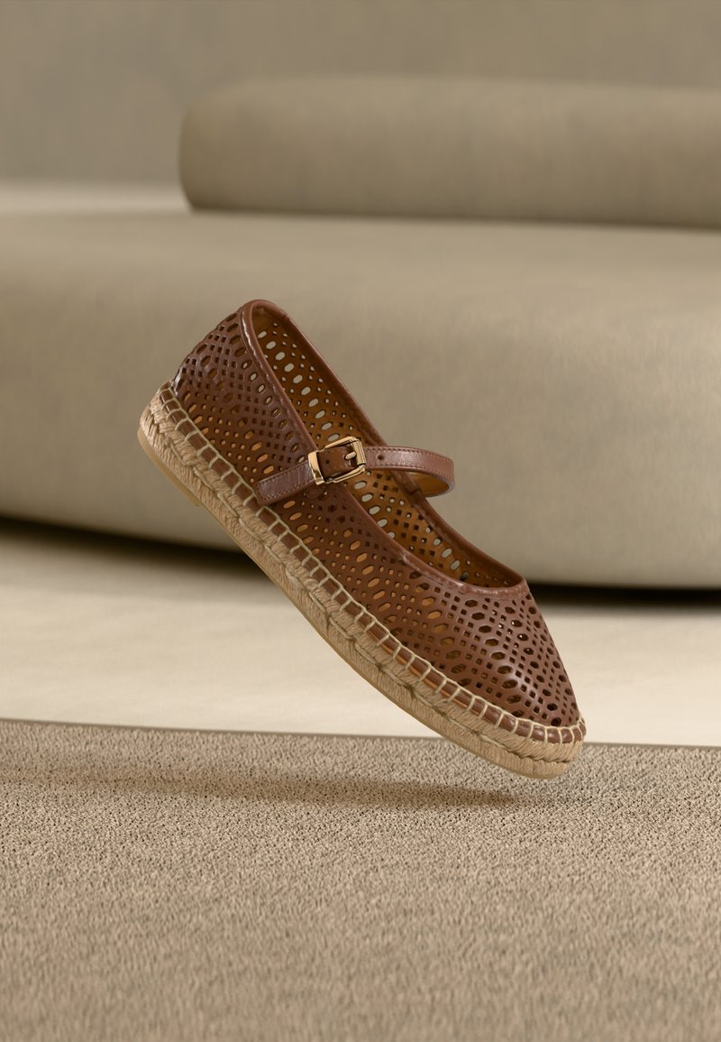 Chaussure en cuir marron perforée avec une bride et une boucle dorée, dotée d'une semelle en jute tressé et d'un bout arrondi. Flottant au-dessus d'une surface texturée.