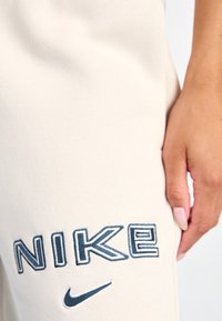 Beige Collegehousereita, jossa on sininen brodeerattu "NIKE"-logo ja viuhkatieto. Sileä, pehmeä kangas ja rento istuvuus. Käsivarsi sijoitettuna kankaalle.