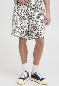 Shorts blancs avec imprimé botanique noir, tissu léger, coupe décontractée, poches latérales, portés avec des baskets noires et des chaussettes blanches mi-hautes.