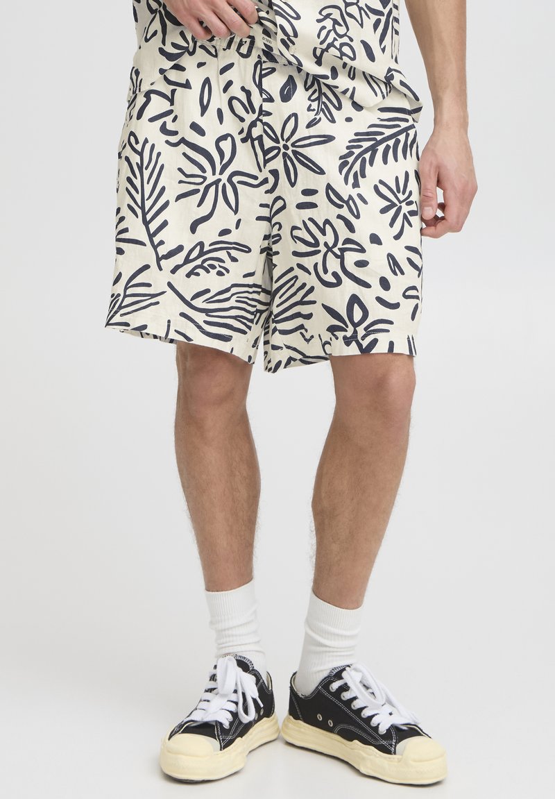 White shorts with black botanical print, lightweight fabric, relaxed fit, side pockets, worn with black sneakers and white crew socks.