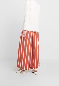 Bílý top s dlouhým rukávem ve spojení se širokými rezavě zbarvenými kalhotami s vertikálními bílými pruhy. Outfit doplňují bílé boty s masivním podpatkem.