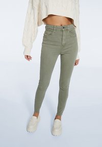 Jeans ajustados de talle alto en verde oliva con bolsillos frontales, combinados con un suéter corto de punto trenzado color crema y mocasines blancos de plataforma.