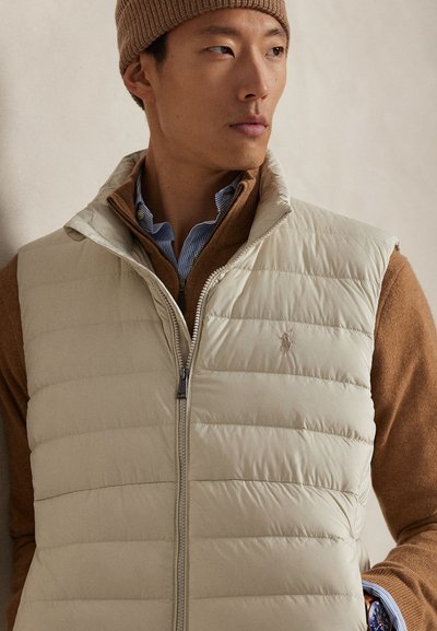 Beige vadderad väst med vertikal quiltad design, dragkedja och logodetalj; bärs över en brun tröja, lager-på-lager med en blå- och vitrandig skjorta.