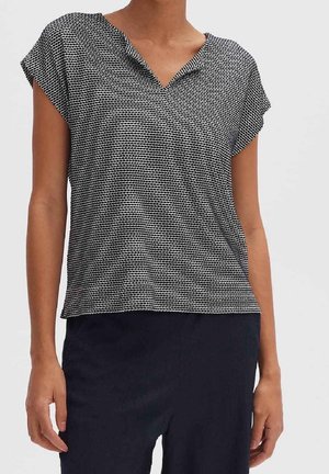 Femme portant un haut à manches courtes noir et blanc à motifs avec un col en V et un pantalon foncé, représentée de la nuque aux hanches sur un fond uni.