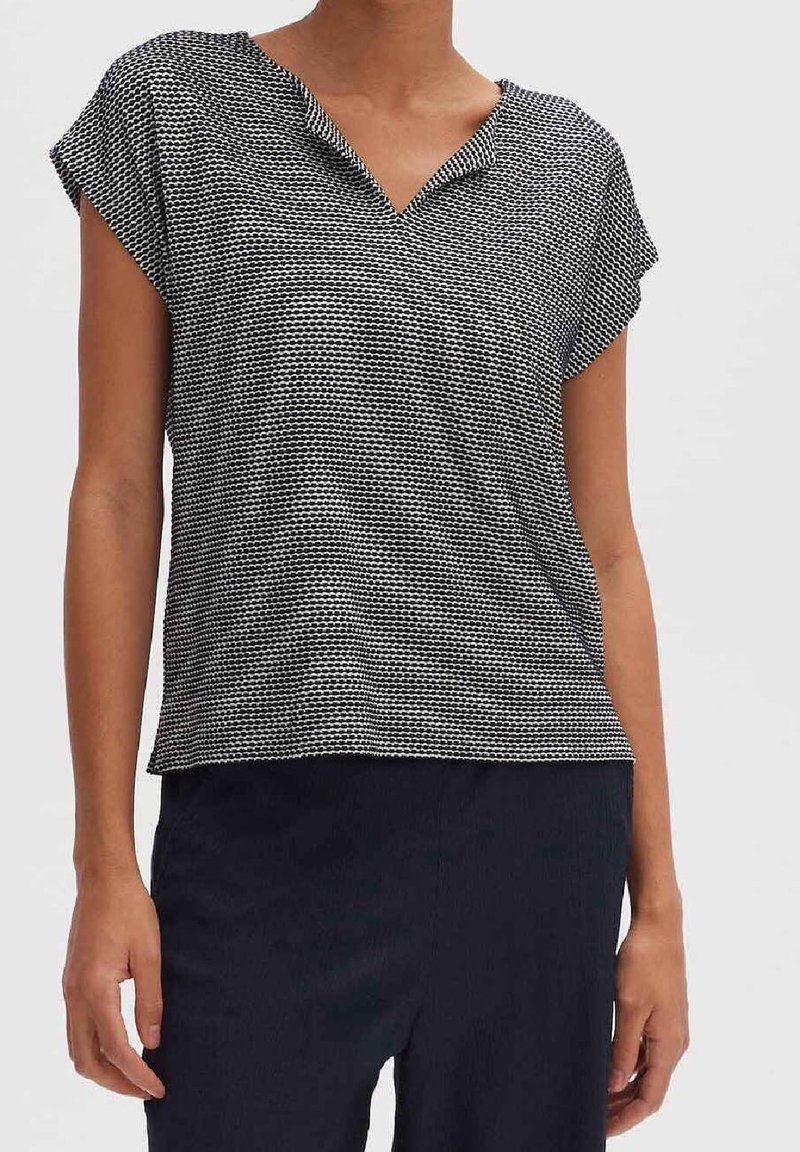 Femme portant un haut à manches courtes noir et blanc à motifs avec un col en V et un pantalon foncé, représentée de la nuque aux hanches sur un fond uni.