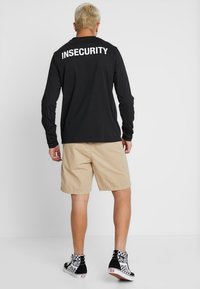 Schwarzes Langarmshirt mit weißem Text "INSECURITY" auf dem Rücken, kombiniert mit beigen Shorts und schwarz-weißen karierten Turnschuhen.