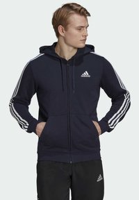 Sudadera con cremallera de color marino con rayas blancas en las mangas, que cuenta con una capucha ajustable, bolsillos frontales y un logo de Adidas en el pecho.