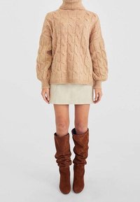 Maglione a trecce spesso di un beige chiaro con colletto alto, abbinato a una minigonna beige e stivali marroni morbidi fino al ginocchio.