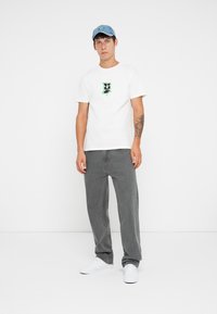 Hvit grafisk T-skjorte med et svart og grønt design, kombinert med grå denimbukser og hvite joggesko. En lyseblå caps fullfører antrekket.