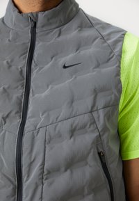 Grå puffväst med hög krage, med svart dragkedja och Nike-logotyp, kombinerad med en neongul, kortärmad, perforerad skjorta under.