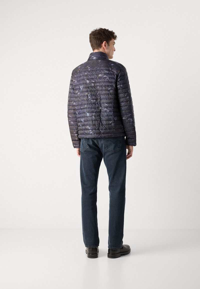 Pikowana kurtka puchowa w ciemnoniebieskim kolorze z błyszczącą powierzchnią, z kołnierzem typu stójka i poziomym przeszyciem. Połączona z ciemnym dżinsami.