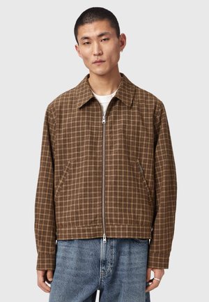 Mand iført brun ternet lynlåstrøje over hvid skjorte og blå jeans, stående mod en ensfarvet lys grå baggrund.