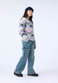 Jersey gris de punto con patrones de texto multicolor, combinado con jeans cargo de color azul claro que tienen bolsillos laterales y piernas acampanadas. Botas negras completan el look.