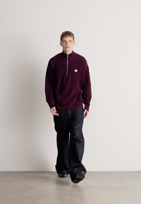 Tumši sarkans adīts džemperis ar pusdaļēju rāvējslēdzēja apkakli, kopā ar tumšām džinsa biksēm. Modelis stāv pret vienkāršu fonu, rokas gar sāniem.