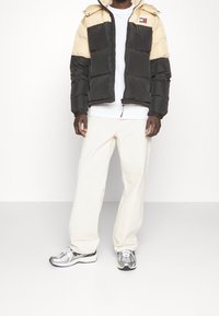 Svart och beige puffjacka med huva, frontdragkedja och logotypmärke. Bärs över en vit skjorta och tillsammans med krämfärgade byxor och silverfärgade sneakers.