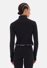 Juodas ilgomis rankovėmis sportinis viršus su aukštu apykakliniu uždaru ir tekstūruotu audiniu. Yra trumpas dizainas ir su ženkline juosta, rodanti prigludusią siluetą.