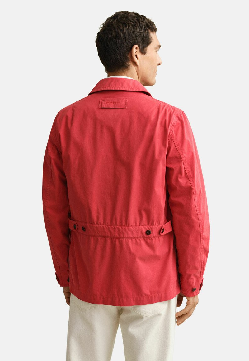 Homme portant une veste rouge à poignets boutonnés avec des pattes de serrage à la taille, vu de dos, associée à un pantalon de couleur claire.