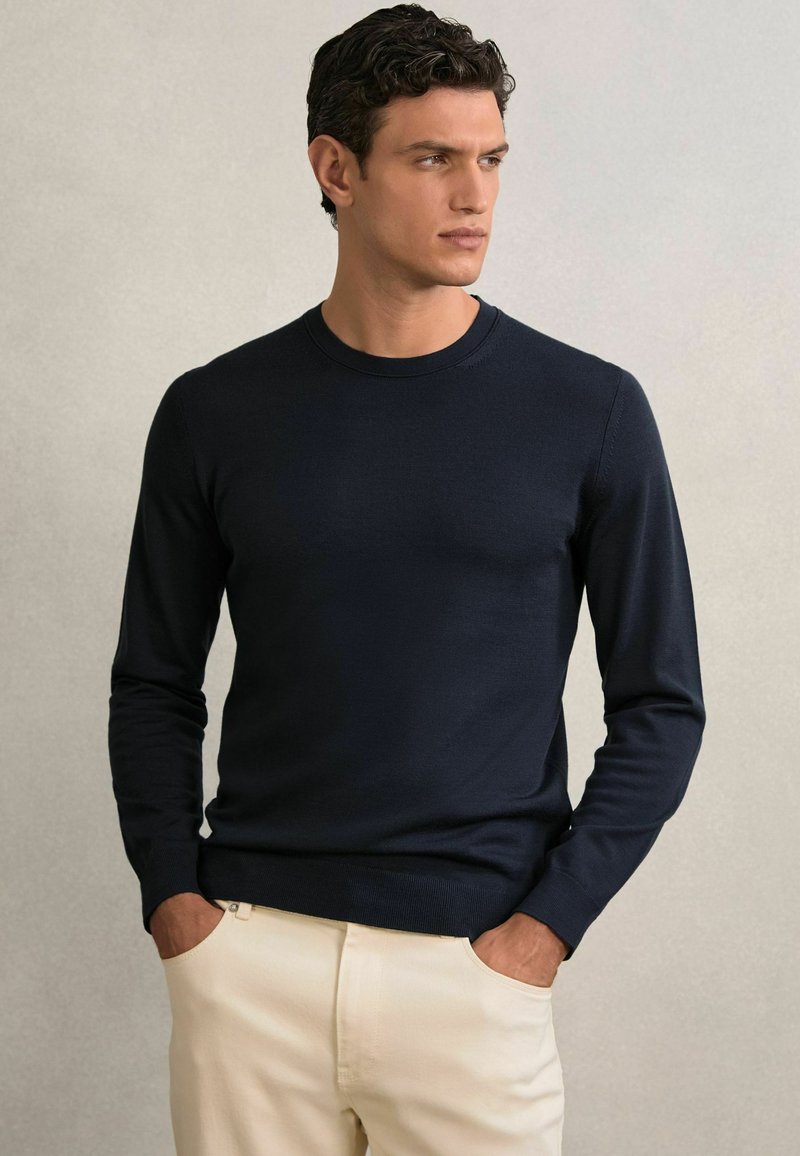 Maglione a maniche lunghe navy a maglia con colletto tondo, texture liscia e polsini aderenti, abbinato a pantaloni chiari.