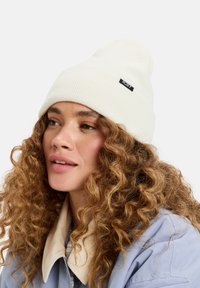 Witte geribde beanie met een omgebogen rand en een zwart Roxy-label. Model heeft krullend haar en draagt een lichtblauwe jas met een beige kraag.