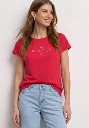 Frau lächelt, trägt ein rotes „Palm Club“ T-Shirt und hellblaue Jeans und steht vor einer weißen strukturierten Wand.