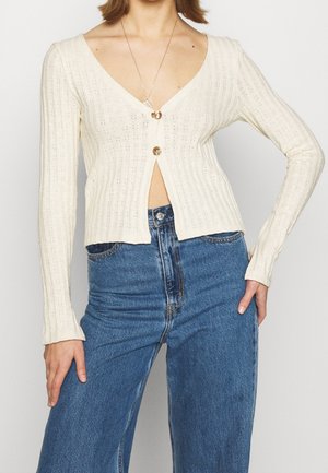 Cream-colored ribbed cardigan with a deep V-neck, featuring two buttons at the front. Paired with high-waisted blue jeans.