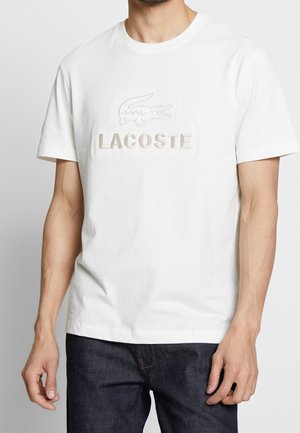 Biała bawełniana koszulka z haftowanym logo krokodyla oraz napisem "LACOSTE" w kolorze beżowym. Klasyczny okrągły dekolt i krótkie rękawy.