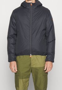 Marineblauer Pufferjacke mit Kapuze, mit einem Frontreißverschluss mit orangefarbener Ziehlasche und elastischen Bündchen. Trägt sie über olivgrünen Cargo-Hosen.