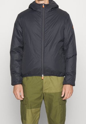 Navy puffer jacket with a hood, featuring a front zipper with orange pull, and elastic cuffs. Worn over olive cargo pants.