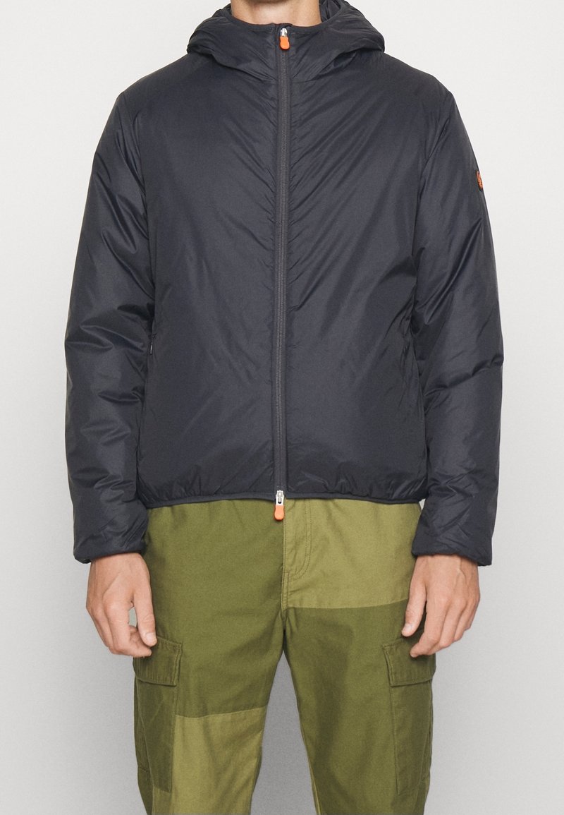 Marineblauer Pufferjacke mit Kapuze, mit einem Frontreißverschluss mit orangefarbener Ziehlasche und elastischen Bündchen. Trägt sie über olivgrünen Cargo-Hosen.
