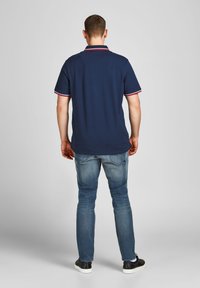 Marineblauwe polo met contrasterende rode en witte kraag en mouwafwerkingen, gecombineerd met lichtblauwe spijkerbroek en zwarte sneakers.