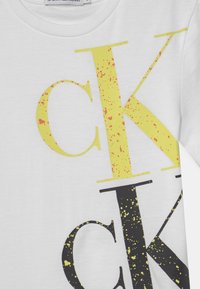 Hvidt stof med store overlappende CK-bogstaver i gul og sort, sprøjtet med kontrasterende malerpletter, og en Calvin Klein Jeans-label ved kraven.