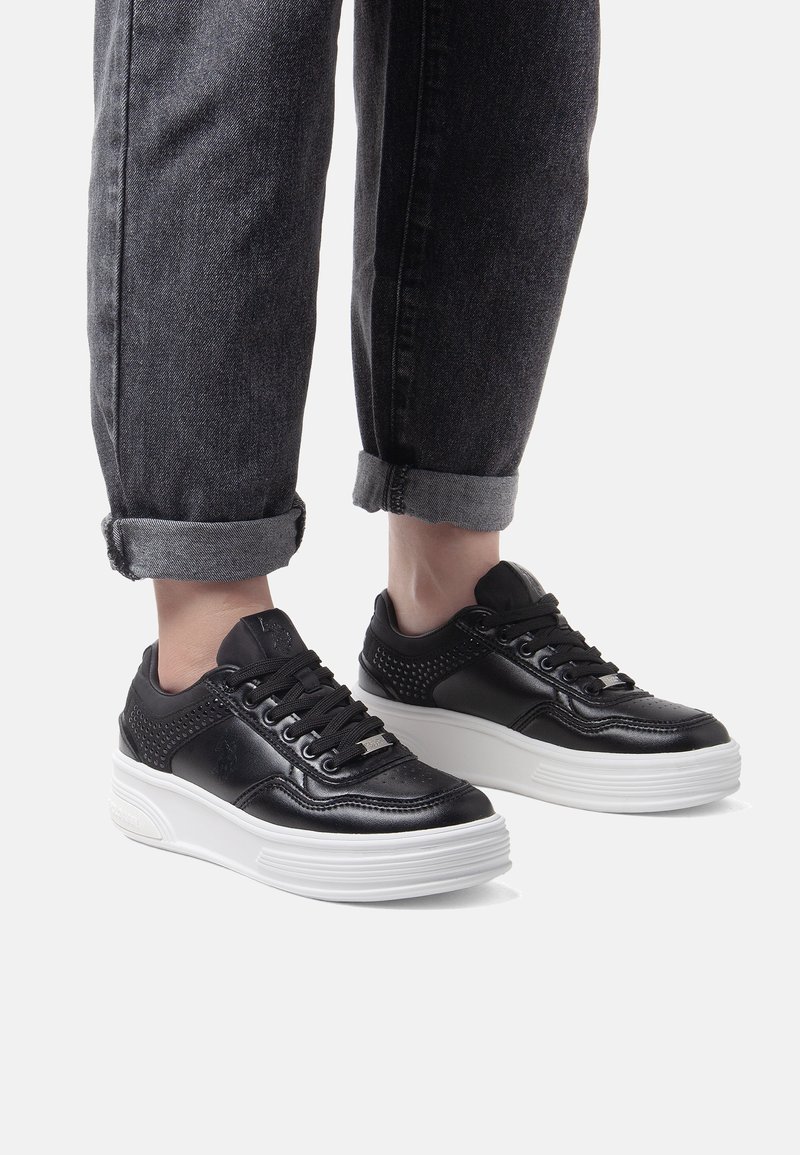 Svarta lädersneakers med perforerade detaljer, vit platt sula, rund tå och svarta snören, tillsammans med uppvikta mörka jeans.